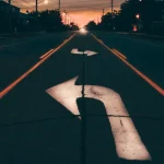 Carretera al atardecer con flechas pintadas en el asfalto señalando distintas direcciones, símbolo de la decisión entre invertir en fondos o ETF