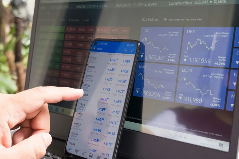 Mano sosteniendo un móvil con una app de cotizaciones en tiempo real, con gráficos de índices bursátiles de fondo, representando la negociación de ETF en bolsa.