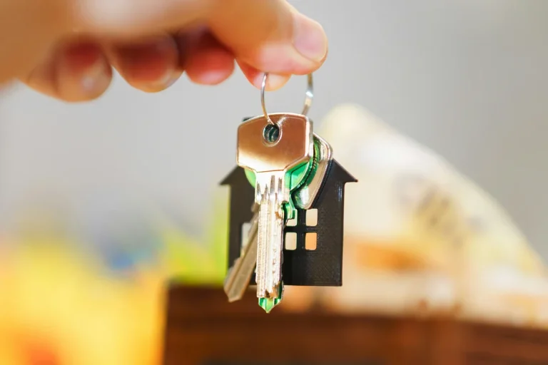 Mano sosteniendo unas llaves con un llavero en forma de casa, con billetes y monedas de fondo, símbolo de la inversión inmobiliaria.