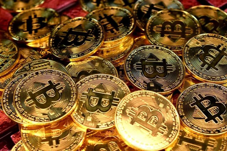Montón de monedas físicas doradas con el símbolo de Bitcoin sobre fondo rojo, representando la criptomoneda.